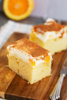 Zwei Stücke Schmandkuchen vom Blech mit Mandarinen auf einem Holzbrett mit angelegtem Messer.