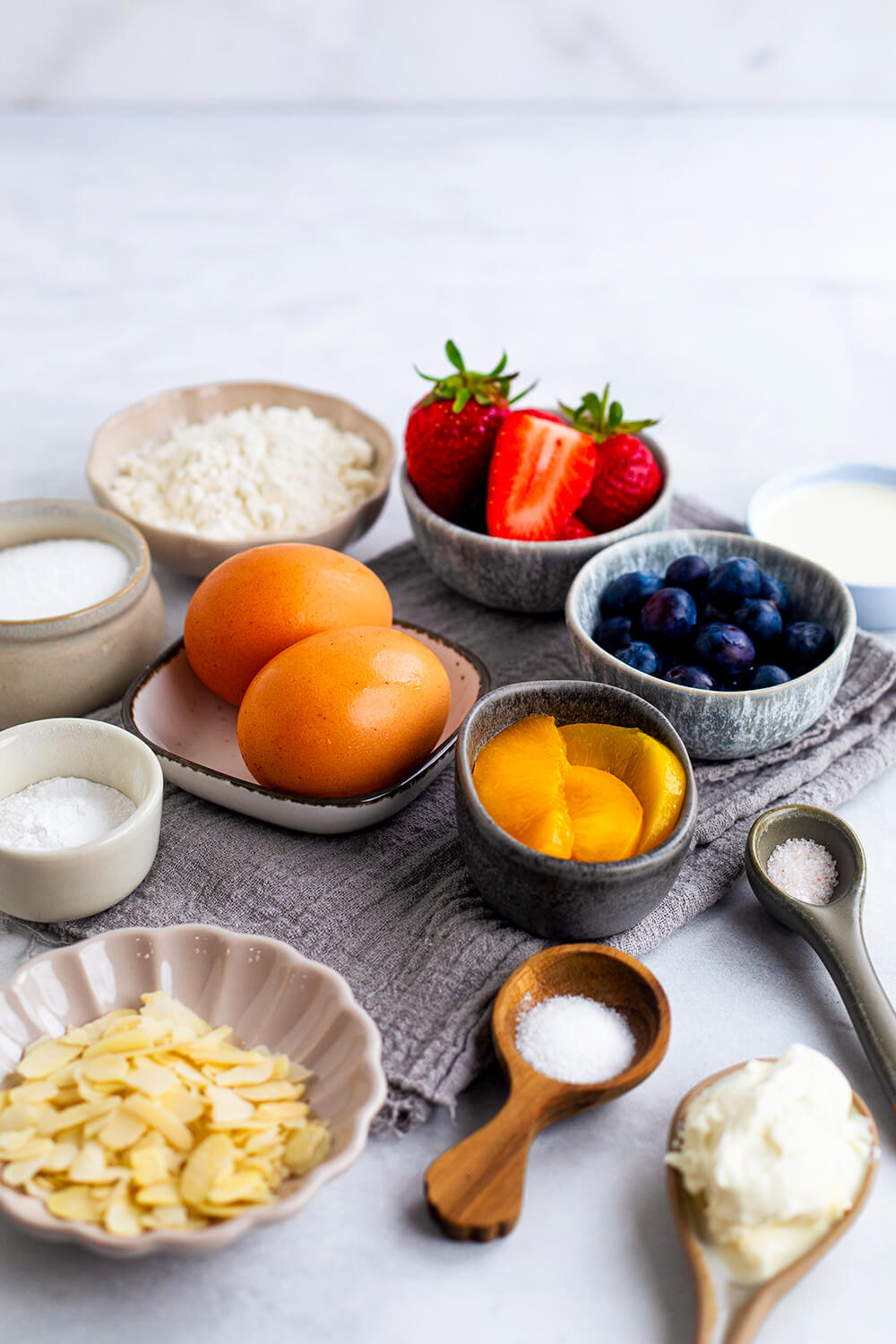 Dreierlei Obst, Eier, Mandelblättchen und Zutaten für den Biskuit in Schälchen angerichtet.