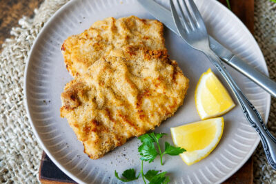 Schnitzel aus der Heißluftfritteuse auf einem Teller mit Zitrone, Petersilie und angelegtem Besteck.