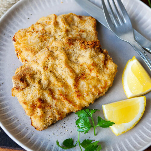 Schnitzel aus der Heißluftfritteuse auf einem Teller mit Zitrone, Petersilie und angelegtem Besteck.